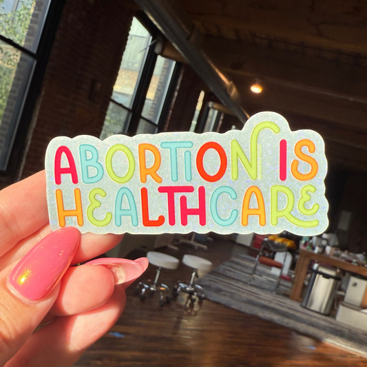 Hand holding a colorful sticker with 'ABORTION IS HEALTHCARE' text in an indoor setting.