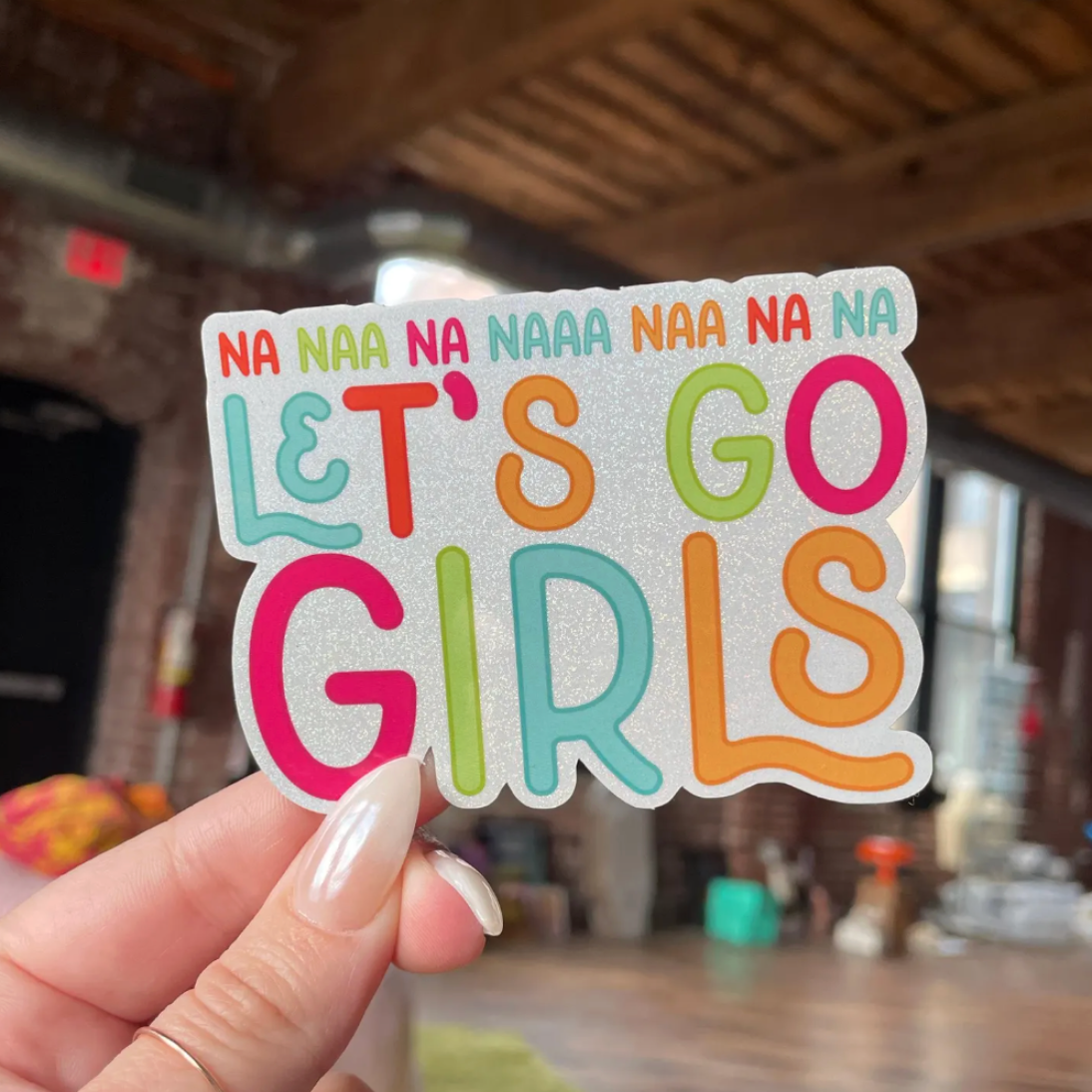 Colorful sticker with 'Let's Go Girls' text held by a hand indoors.