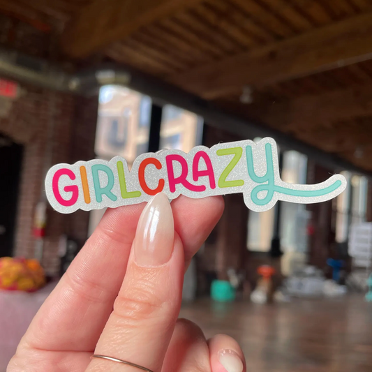 Hand holding a colorful 'GIRLCRAZY' sticker with a blurred indoor background.