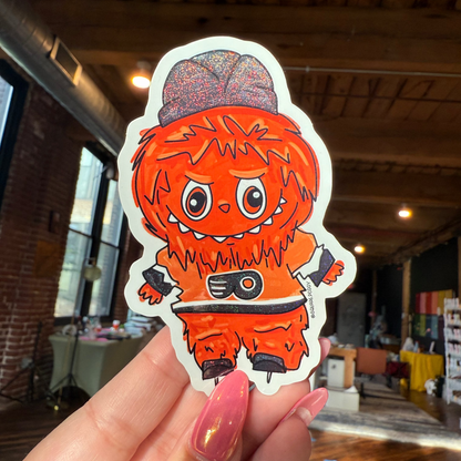 Hand holding a sticker of an orange character with a hat in a casual indoor setting