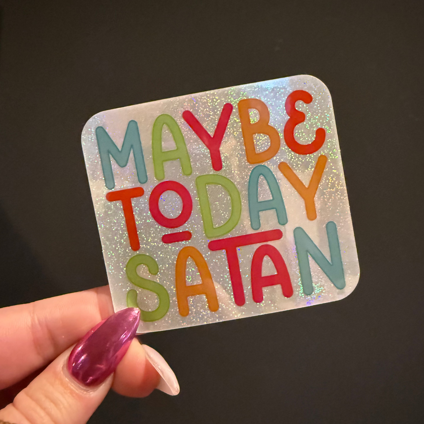 Holographic sticker with colorful text 'MAYBE TODAY SATAN' held by a hand against a dark background