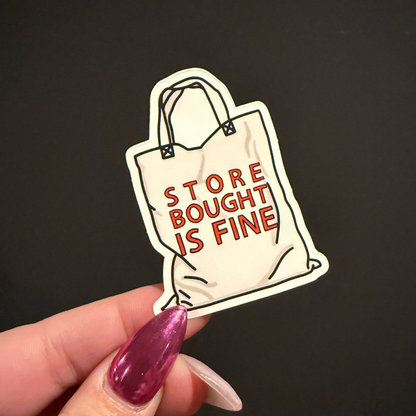 Sticker of a shopping bag with 'Store Bought is Fine' text held by a hand against a black background