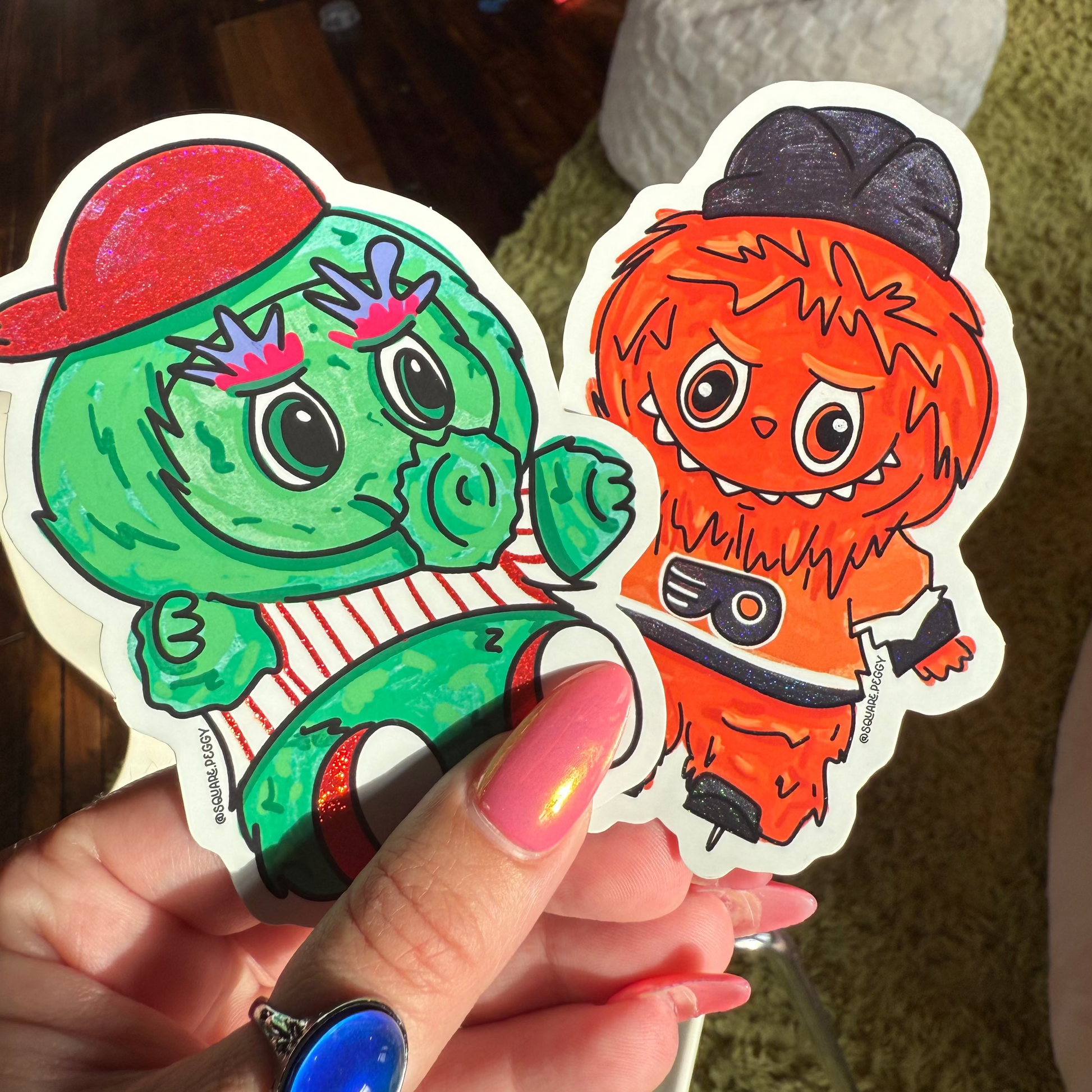 Two colorful stickers of cartoon characters held by a person with a ring.