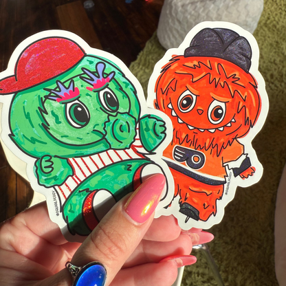 Two colorful stickers of cartoon characters held by a person with a ring.